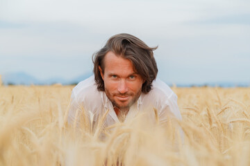 The handsome brutal young man with long brunette hairs poses in the field with wheat, suntanned...
