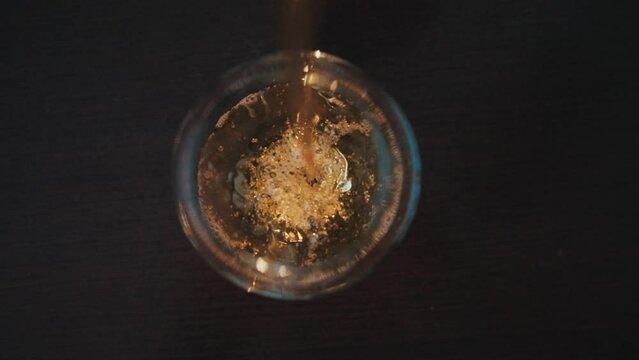 A brown liquid is poured into glass beaker. Alcoholic beverage is poured into vessel and bubbles are formed. Top view. Nightlife at bar. The bartender pours whiskey from bottle. Relaxation with booze