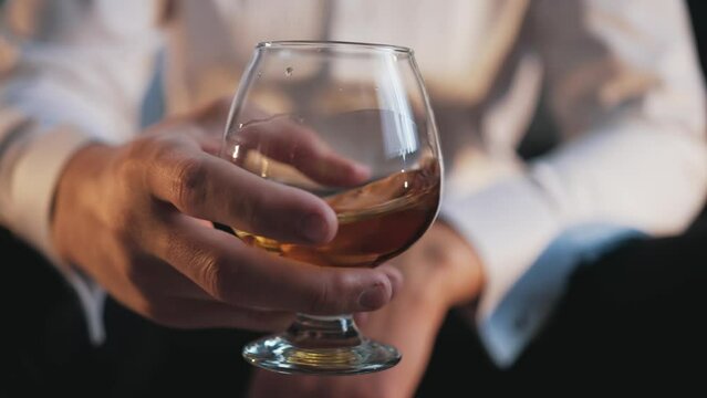 The man sits alone in the room and holds a glass of rum in his hand and shakes glass so that the alcohol flows down the walls of the vessel. Upset man drinking alcohol. Relax with alcohol. Alcoholism