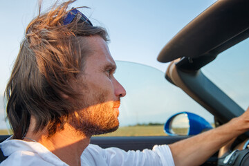 Fototapeta na wymiar The handsome brutal man with long hair and an easy beard at a car without top at sunset, cabriolet, blue color, sunglasses