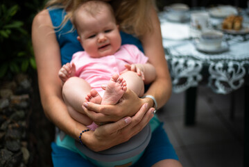 mother holds baby girl, mother's hands holding little baby feet, garden table with porcelain tableware in a blurred background