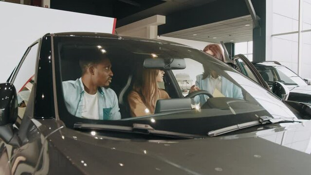 Beautiful Multiracial Couple Sitting Inside Modern Luxury Car And Examining Details Of Interior. Family Choosing Vehicle Together At Auto Salon. Saleswoman Showing To Diverse Couple Car At Showroom.