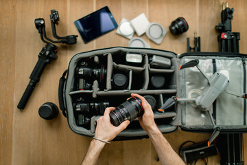 Professional  photographer packs smart backpack with equipment, holding  lens. Social media influencer top view on suitcase with lens, videocamera and camera, amateur drone and gimbal