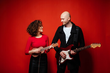 Curly cheerful girl in a red dress plays ukulele. Bald dude playing red guitar.