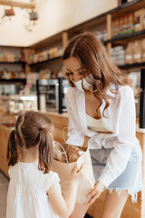 A young mother and daughter in medical masks are shopping with a paper bag at the counter in cafe or small shop. Concept of a retail business during a pandemic