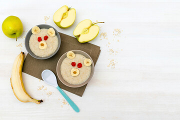 Funny cute kids childrens baby's healthy breakfast lunch oatmeal porridge in bowl look like bear face decorated with apple, banana, dried berry fruits. dessert food art on white wooden table