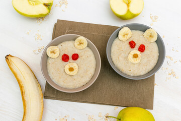 Fototapeta na wymiar Funny cute kids childrens baby's healthy breakfast lunch oatmeal porridge in bowl look like bear face decorated with apple, banana, dried berry fruits. dessert food art on white wooden table