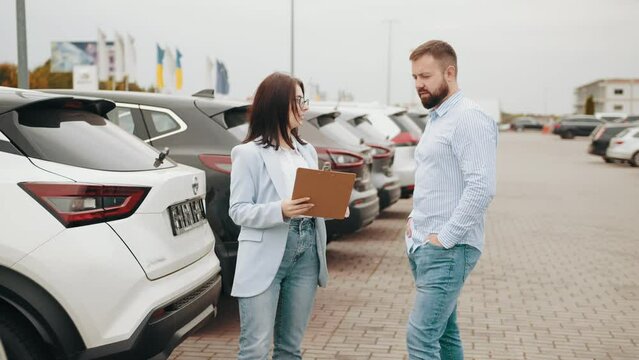 Male customer having conversation with competent saleswoman while choosing car at showroom. Man and woman standing outdoors near row of luxury autos. Female seller helping male client shoosing car.