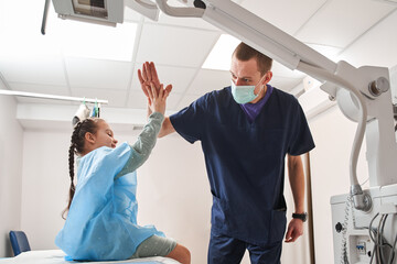 Male doctor giving five to the little child girl in a hospital bed after successful radiotherapy