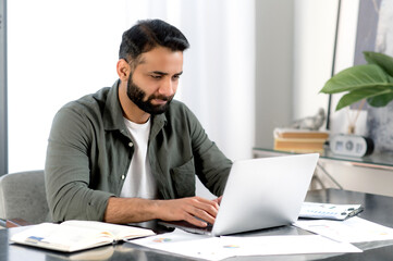 Fototapeta premium Concentrated confident indian man using laptop in modern office. Successful businessman, freelancer, manager, in casual shirt, browsing email, working and typing on laptop, working on a project