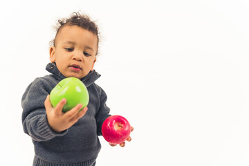 Fototapeta na wymiar Biracial baby boy with curly hair looking at juicy green apple in one hand and holding red apple in the other. Healthy food concept. High quality photo