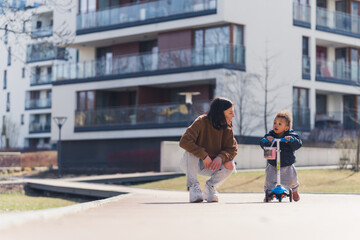 Weekend in the city. Young dedicated mother kneeling down and supporting her biracial little toddler boy who uses a scooter for the first time. High quality photo - Powered by Adobe
