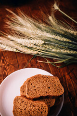 Slices of rye wholegrain bread on white plate, wheat ears on wooden boards background, rustic kitchen table. Homemade bread, baking