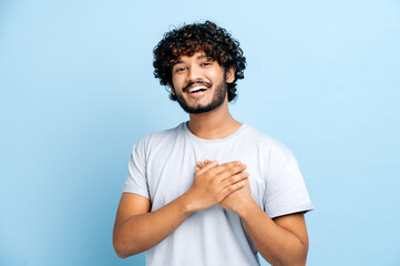 A gesture of sincerity and trust. Handsome candid indian or arabian guy with hands on heart and grateful gesture, smile on face, looks at camera, stand on isolated blue background. Peaceful concept - obrazy, fototapety, plakaty