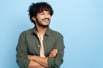 Handsome charismatic successful indian or arabian curly-haired guy in stylish clothes, standing with crossed arms over isolated blue background, looking away, smiling positively