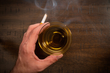 A male caucasian hand holds a smoking cigarette near a glass of whiskey.