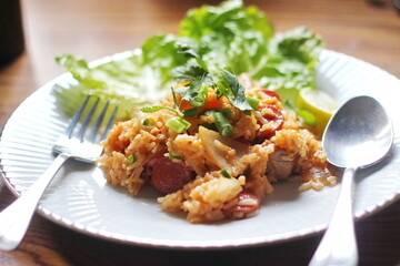 Pork and sausage fried rice served in a white plate on a light wooden table. With lemon, vegetables and tomato slices on a light wooden table.
