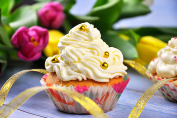 A festive cupcake with white cream and flowers in the background.