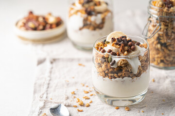 Crunchy granola with yogurt, banana, nuts, chocolate and honey in a glass on white background. Healthy breakfast concept.