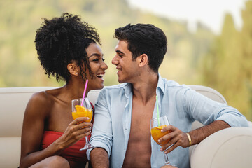Happy biracial couple of young people sit outdoors in a sofa in the garden looking at each other while drinking healthy fruit cocktails - love and vacation lifestyle concept - Powered by Adobe