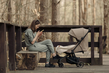 The young mom is working with her cellphone while her infant child is sleeping in the baby stroller. The mother with her child in the park for fresh air.