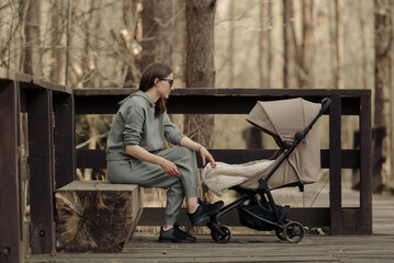 The young mom is relaxing on the wooden bench while her infant child is sleeping in the baby stroller. The mother with her child in the park for fresh air.
