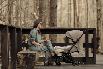 The young mom is relaxing on the wooden bench while her infant child is sleeping in the baby stroller. The mother with her child in the park for fresh air.