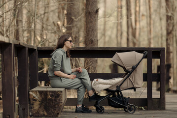 The young mom is relaxing on the wooden bench while her infant child is sleeping in the baby...