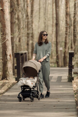 The young mom is walking on the wooden walkway with her infant child who is sleeping in the baby stroller. The mother with her child in the park for fresh air.