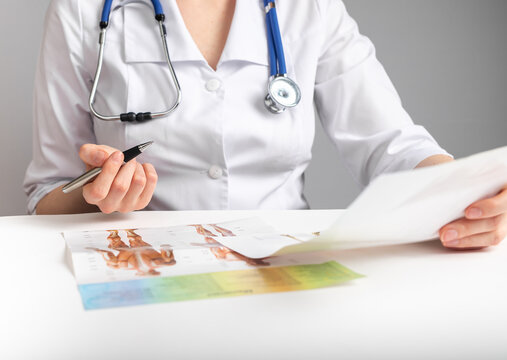 Doctor holding papers and studying anatomy. Medicine education and research conducting concept. Woman in lab coat with stethoscope sitting at table and analyzing documents. High quality photo