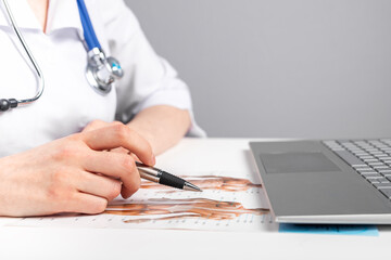 Doctor studying or teaching anatomy. Medicine education, online courses concept. Woman in lab coat with stethoscope sitting at table with laptop and looking at papers with human body image. photo