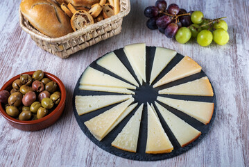 Spanish cheese board cut into wedges and presented on a rustic stone plate, accompanied by grapes, olives and bread.