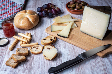 Cutting board with a piece of cheese, accompanied by grapes, jam, olives and assorted bread.