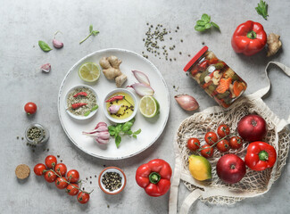 Vegetables preserving preparation with flavoring herbs and spices on grey concrete kitchen table. Oil, tomatoes, bell pepper, garlic, onions and lime. Preserved vegetables in glass jar. Top view.