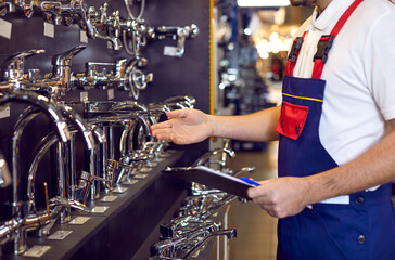 Choice of water taps and faucets. Male worker checks assortment on showcase with stainless steel water taps in plumbing store. Cropped image of salesman in coveralls with clipboard near various taps.