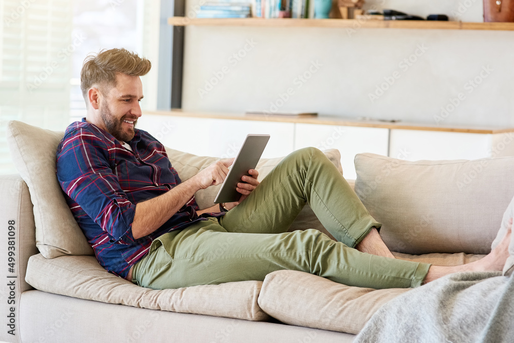 Poster chilled vibes online. shot of a relaxed young man using a digital tablet on the sofa at home.