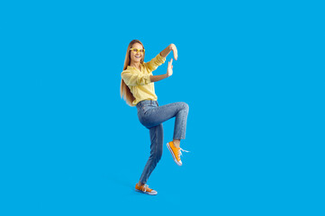 Fototapeta na wymiar Smiling young woman in casual wear and glasses isolated on blue studio background make eccentric funny dancer moves. Happy millennial girl have fun dance enjoy hobby activity. Full length.