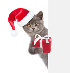 Funny kitte wearing red christmas hat holds gift box behind empty white banner. isolated on white background