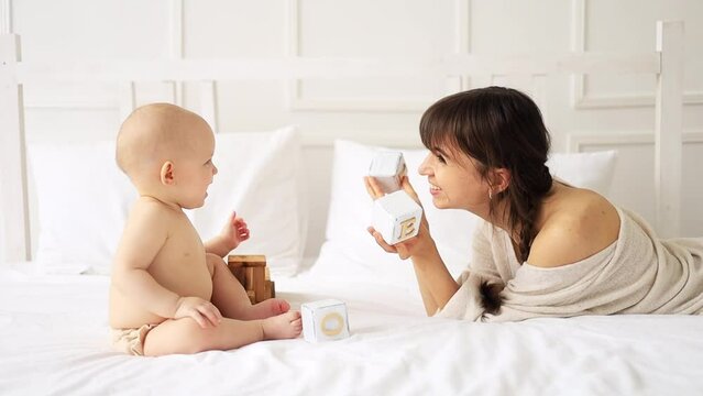 mom with a baby in diapers playing with wooden toys closing her eyes at home on a bed in a bright white room, maternal love and care