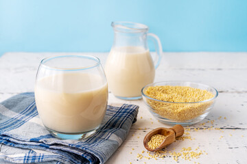 Millet milk in glass, glass jar and bowl on white wooden table with blue background