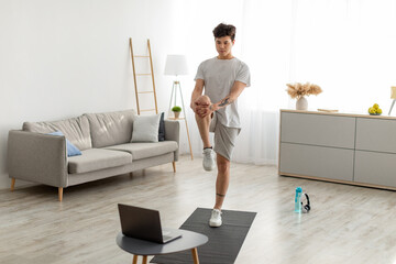 Asian guy doing standing knee to chest exercise using laptop