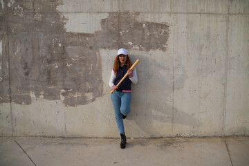 young and beautiful redhead woman is happy with baseball cap, jacket, baseball bat and jeans, she...