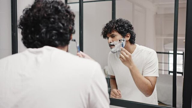 Young Indian man shaves beard with razor looking in mirror
