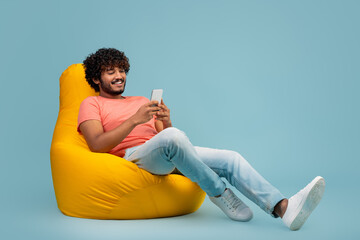 Stylish hindu guy sitting on bean bag, using cell phone