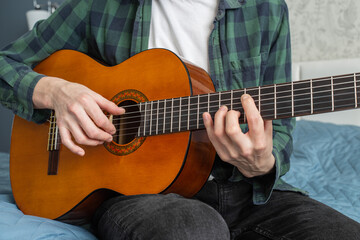 Fototapeta na wymiar A man in a plaid shirt plays a natural-colored classical guitar sitting on a bed in close-up, selective focus. A male musician plays the guitar. Classical guitar in natural color.