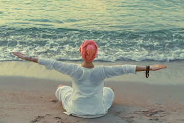 A woman in white clothes and a turban does yoga exercises sitting on the sand by the sea on the...