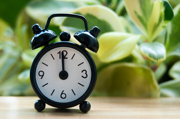 Black alarm clock isolated on blurred nature background. The clock set at 12 o'clock.