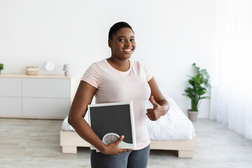 Smiling plus size black woman holding scales, showing thumb up gesture, happy with result of her slimming diet at home