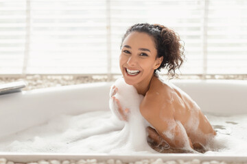 Charming joyful young woman sitting in hot bubble bath covered in foam, relaxing in bathtub at...
