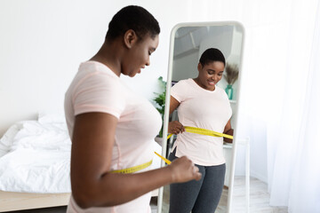 Plus size black woman measuring waist with tape, standing near mirror, satisfied with results of...